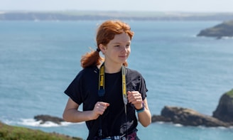 A person with red hair is standing near a coastal area, wearing a black t-shirt and holding a Nikon camera around their neck. The background features a calm sea with distant rocky cliffs and a clear sky.