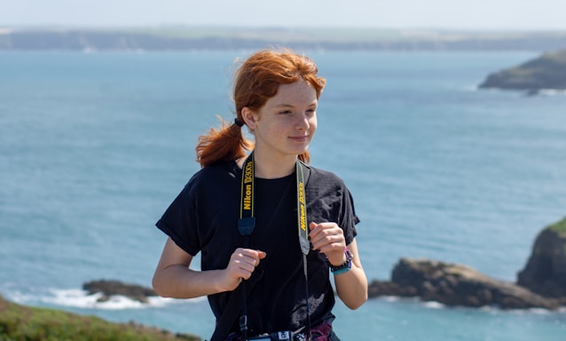 A person with red hair is standing near a coastal area, wearing a black t-shirt and holding a Nikon camera around their neck. The background features a calm sea with distant rocky cliffs and a clear sky.