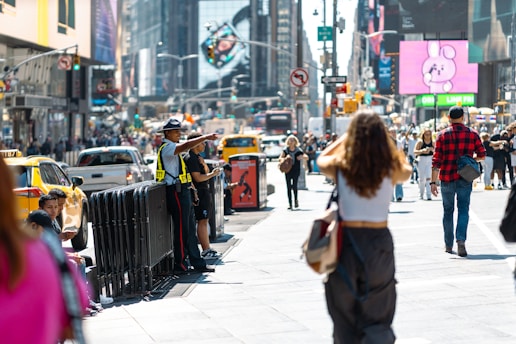 A bustling city street filled with people walking in various directions. A security officer in a hat and reflective vest is directing pedestrians and vehicles. Numerous yellow taxis and other cars populate the street. Large electronic billboards and signs are visible in the background, showcasing advertisements and images.