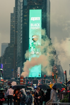 A bustling urban scene with a large digital billboard displaying 'Black Friday' and a character, surrounded by tall buildings. Smoke or steam rises in the foreground while people with umbrellas navigate through a city street filled with traffic and vibrant lights.