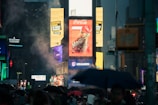 Bright electronic billboards illuminate a busy cityscape under a cloudy, possibly rainy sky. The captivating advertisements include a prominent Coca-Cola sign. Crowds are bustling below, many carrying umbrellas, suggesting a drizzly environment.