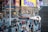 A busy urban street scene with numerous pedestrians walking past various shops and advertisements. Bright, colorful billboards display promotional content for musicals and artists. In the center, an FDNY ambulance is visible among the traffic. The area is bustling with activity, indicative of a popular city location, possibly a tourist spot, with diverse people engaged in various activities.