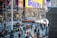 A busy urban street scene with numerous pedestrians walking past various shops and advertisements. Bright, colorful billboards display promotional content for musicals and artists. In the center, an FDNY ambulance is visible among the traffic. The area is bustling with activity, indicative of a popular city location, possibly a tourist spot, with diverse people engaged in various activities.