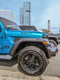 A bright blue Jeep Wrangler with 'Willys' written on its side is parked in an urban area. The vehicle's large black tires are clearly visible. Modern high-rise buildings stand in the background under a clear blue sky.