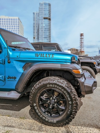 A bright blue Jeep Wrangler with 'Willys' written on its side is parked in an urban area. The vehicle's large black tires are clearly visible. Modern high-rise buildings stand in the background under a clear blue sky.