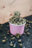 A purple paper cup filled with green cannabis buds is placed on a dark surface. Additional buds are scattered around the cup, and a light wooden background is visible.