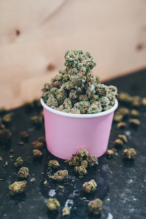 A purple paper cup filled with green cannabis buds is placed on a dark surface. Additional buds are scattered around the cup, and a light wooden background is visible.