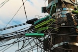 A wooden utility pole with numerous tangled cables and wires is visible. The cables are held together by green plastic clips and are clearly interconnected with various metal fittings and insulators. The sky forms a soft blue backdrop, giving the scene a slightly chaotic yet industrial appearance.