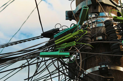 A wooden utility pole with numerous tangled cables and wires is visible. The cables are held together by green plastic clips and are clearly interconnected with various metal fittings and insulators. The sky forms a soft blue backdrop, giving the scene a slightly chaotic yet industrial appearance.