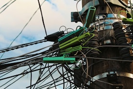 A wooden utility pole with numerous tangled cables and wires is visible. The cables are held together by green plastic clips and are clearly interconnected with various metal fittings and insulators. The sky forms a soft blue backdrop, giving the scene a slightly chaotic yet industrial appearance.