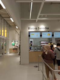 A modern bistro setting with a minimalist design features a counter with menu boards displaying food items and prices. Several people stand in front of the counter, possibly ordering. The interior has a bright and clean look with tile accents and neutral colors.