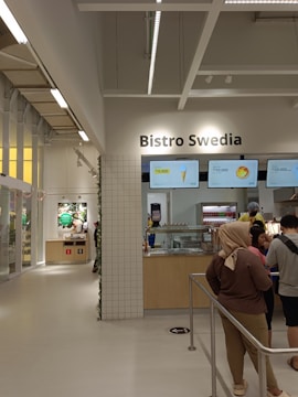 A modern bistro setting with a minimalist design features a counter with menu boards displaying food items and prices. Several people stand in front of the counter, possibly ordering. The interior has a bright and clean look with tile accents and neutral colors.