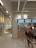 A modern bistro setting with a minimalist design features a counter with menu boards displaying food items and prices. Several people stand in front of the counter, possibly ordering. The interior has a bright and clean look with tile accents and neutral colors.