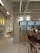 A modern bistro setting with a minimalist design features a counter with menu boards displaying food items and prices. Several people stand in front of the counter, possibly ordering. The interior has a bright and clean look with tile accents and neutral colors.