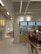 A modern bistro setting with a minimalist design features a counter with menu boards displaying food items and prices. Several people stand in front of the counter, possibly ordering. The interior has a bright and clean look with tile accents and neutral colors.