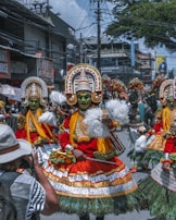 A vibrant street parade features elaborately dressed performers in traditional costumes with colorful face paint and ornate headdresses. These individuals are engaged in a cultural dance, carrying pom-poms and surrounded by onlookers and a photographer capturing the moment. The backdrop includes urban elements like power lines, buildings, and signage.