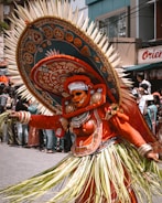 A performer in intricate traditional attire participates in a cultural festival. The costume is vibrant with detailed patterns and embellishments, featuring a large, ornate headdress with spikes and rich embroidery. The outfit includes a combination of orange fabric and natural materials, and the performer's face is painted elaborately, adding to the dramatic effect. Numerous spectators are visible in the background, suggesting a lively public event.