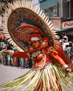 A performer in intricate traditional attire participates in a cultural festival. The costume is vibrant with detailed patterns and embellishments, featuring a large, ornate headdress with spikes and rich embroidery. The outfit includes a combination of orange fabric and natural materials, and the performer's face is painted elaborately, adding to the dramatic effect. Numerous spectators are visible in the background, suggesting a lively public event.