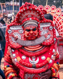 A person dressed in traditional, elaborate ceremonial attire dominated by intricate patterns and vibrant colors, primarily red with silver accents. The costume includes a large, ornate headdress and facial makeup, suggesting a cultural or religious festival setting. Other people and similar costumes are visible in the background.