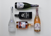 Five bottles of various alcoholic beverages are arranged neatly on a light background. The bottles include a white frosted glass vodka bottle, a black rectangular gin bottle, a brown glass whiskey bottle, and two bottles of sparkling wine with decorative labels.