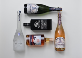 Five bottles of various alcoholic beverages are arranged neatly on a light background. The bottles include a white frosted glass vodka bottle, a black rectangular gin bottle, a brown glass whiskey bottle, and two bottles of sparkling wine with decorative labels.