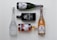 Five bottles of various alcoholic beverages are arranged neatly on a light background. The bottles include a white frosted glass vodka bottle, a black rectangular gin bottle, a brown glass whiskey bottle, and two bottles of sparkling wine with decorative labels.
