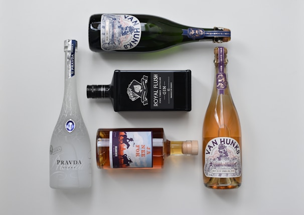 Five bottles of various alcoholic beverages are arranged neatly on a light background. The bottles include a white frosted glass vodka bottle, a black rectangular gin bottle, a brown glass whiskey bottle, and two bottles of sparkling wine with decorative labels.