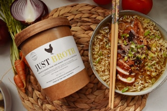 A round bowl filled with ramen noodles topped with sliced roasted meat, greens, and garnishes is placed on a woven mat. Next to it, there is a brown paper container labeled 'Just Broth' with illustrations and text indicating the product is chicken broth from Cape Town, South Africa. On the side lies an arrangement of fresh vegetables including a halved red onion and carrots.
