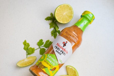 A bottle of Nando's PERi-PERi sauce lies on a light surface, surrounded by fresh lime slices and sprigs of cilantro. The sauce label indicates it is 'Lemon & Herb' flavor and 'extra mild'. The vibrant green, yellow, and brown colors on the label complement the natural green of the lime and cilantro.