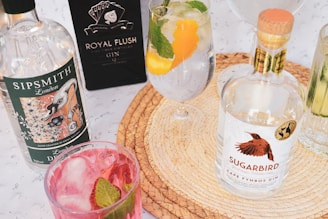 An assortment of gin bottles and cocktails is arranged on a woven placemat. Sipsmith and Sugarbird gin bottles are prominently displayed alongside glasses filled with colorful cocktails garnished with fruit and mint leaves.
