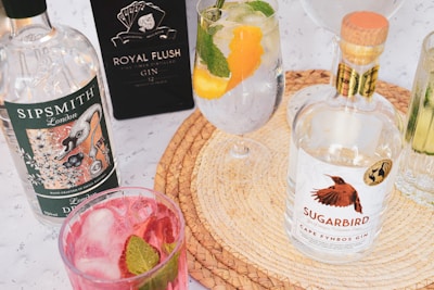 An assortment of gin bottles and cocktails is arranged on a woven placemat. Sipsmith and Sugarbird gin bottles are prominently displayed alongside glasses filled with colorful cocktails garnished with fruit and mint leaves.
