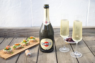 A large bottle of Martini Prosecco is placed on a wooden surface next to two filled champagne flutes. To the left, a wooden board holds several pieces of bread topped with a spread, leafy greens, and cherry tomato halves. In the background, there is a small bowl filled with dark olives.