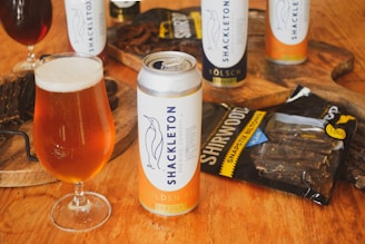 A can of Shackleton Golden Ale and a filled glass are positioned prominently on a wooden surface. In the background, additional cans and a bag labeled 'Shirwood' containing biltong are visible, accompanied by a wooden board with sliced biltong.