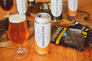 A can of Shackleton Golden Ale and a filled glass are positioned prominently on a wooden surface. In the background, additional cans and a bag labeled 'Shirwood' containing biltong are visible, accompanied by a wooden board with sliced biltong.