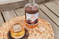 A bottle of liquor with a label featuring silhouettes of cowboys on horseback is placed on a woven mat. Next to the bottle is a glass containing a drink with a large ice cube and a wooden stopper lying on the mat. The setting is on a wooden outdoor surface.