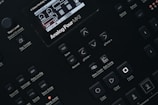 Close-up of a digital synthesizer control panel with various buttons and a small display screen showing audio parameters. The layout features buttons for saving, editing, and managing sound settings with labels like 'Analog Four MKII', 'Tap Tempo', and 'Edit Song'.