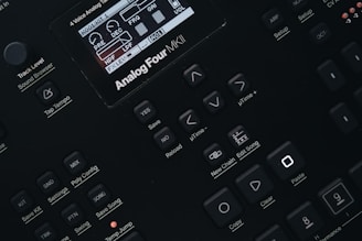 Close-up of a digital synthesizer control panel with various buttons and a small display screen showing audio parameters. The layout features buttons for saving, editing, and managing sound settings with labels like 'Analog Four MKII', 'Tap Tempo', and 'Edit Song'.