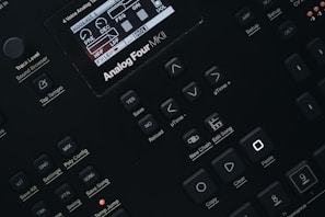 Close-up of a digital synthesizer control panel with various buttons and a small display screen showing audio parameters. The layout features buttons for saving, editing, and managing sound settings with labels like 'Analog Four MKII', 'Tap Tempo', and 'Edit Song'.