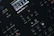 Close-up of a digital synthesizer control panel with various buttons and a small display screen showing audio parameters. The layout features buttons for saving, editing, and managing sound settings with labels like 'Analog Four MKII', 'Tap Tempo', and 'Edit Song'.