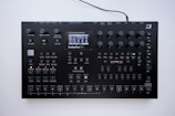 A black electronic music synthesizer with numerous knobs, buttons, and a small display screen in the center. It features labeled controls for various functions like volume, tempo, and different sound effects. The device appears to be designed for complex audio manipulation and production.