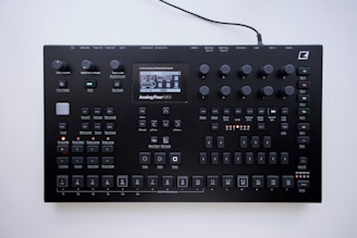 A black electronic music synthesizer with numerous knobs, buttons, and a small display screen in the center. It features labeled controls for various functions like volume, tempo, and different sound effects. The device appears to be designed for complex audio manipulation and production.