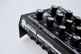 A close-up view of an analog bass synthesizer with multiple black knobs and detailed white labeling. The device features various controls for audio output and input, pitch, filter, and volume adjustments. The brand name 'MINITAUR' is prominently displayed, along with graphical design elements.