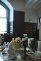 A dining room table is set with a lace tablecloth and various elegant tableware, including teapots, cups, and saucers. A decorative centerpiece with colorful flowers is placed in the middle. A black candelabrum holding white candles is prominently featured. The room has large windows with curtains, and the light filters softly creating a warm, inviting atmosphere.