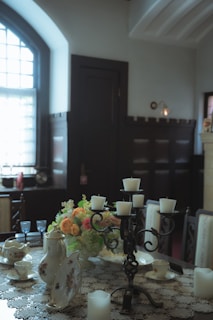 A dining room table is set with a lace tablecloth and various elegant tableware, including teapots, cups, and saucers. A decorative centerpiece with colorful flowers is placed in the middle. A black candelabrum holding white candles is prominently featured. The room has large windows with curtains, and the light filters softly creating a warm, inviting atmosphere.