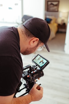 A person wearing a black cap and glasses is operating a professional camera indoors. The camera display shows an image being captured, and the background includes blurred elements like a couch and a framed picture.