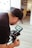 A person wearing a black cap and glasses is operating a professional camera indoors. The camera display shows an image being captured, and the background includes blurred elements like a couch and a framed picture.