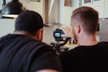 Two individuals are focused on a camera setup, possibly adjusting camera settings or framing a shot. They appear to be collaborating or instructing each other in a well-lit room with white cabinetry and modern fixtures.