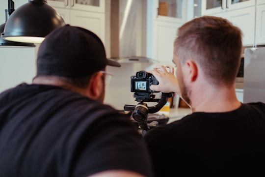 Two individuals are focused on a camera setup, possibly adjusting camera settings or framing a shot. They appear to be collaborating or instructing each other in a well-lit room with white cabinetry and modern fixtures.