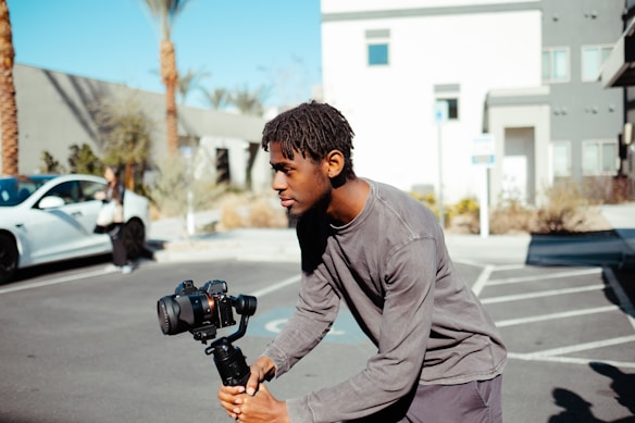 A person is holding a camera mounted on a stabilizer while standing in a parking lot with modern buildings and palm trees in the background.