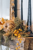 A rustic autumn-themed decoration featuring a pumpkin made of dried leaves and surrounded by various greenery and yellow flowers. Two tall black candles are positioned on a wooden shelf next to an ornate gold mirror frame.